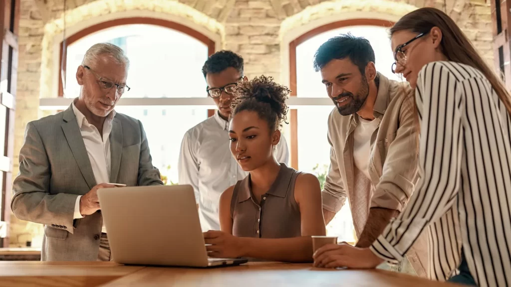 A diverse team comprised of five individuals is intently looking at the screen of a laptop. They seem to be in discussion.