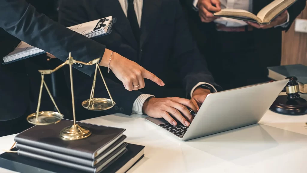 The profiles of what seems to be three lawyers are in a discussion over something one of them is pointing to in a laptop screen. 