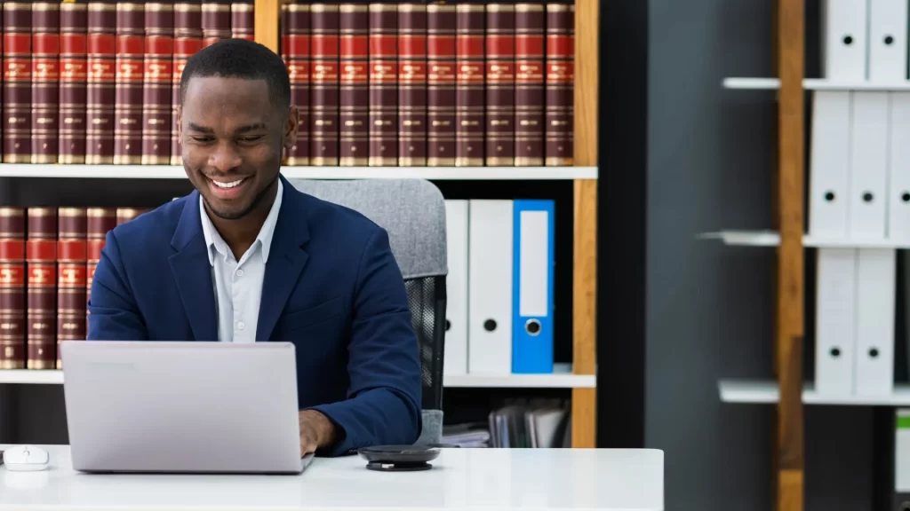 A male lawyer seated in a law library is smiling at something on his laptop.