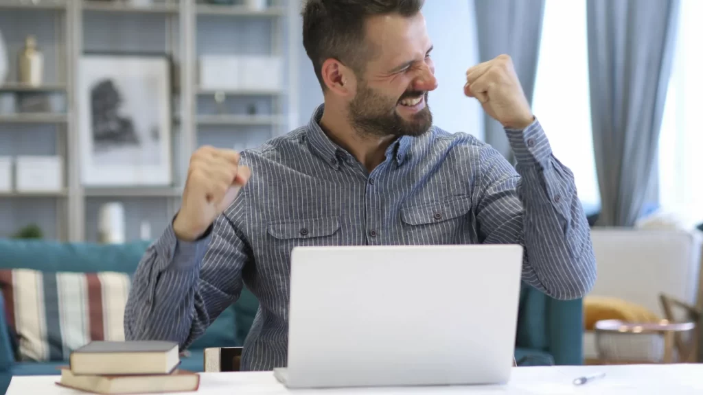 A man in a striped long-sleeved shirt has his arms and fists happily raised while grinning ear to ear in celebration.