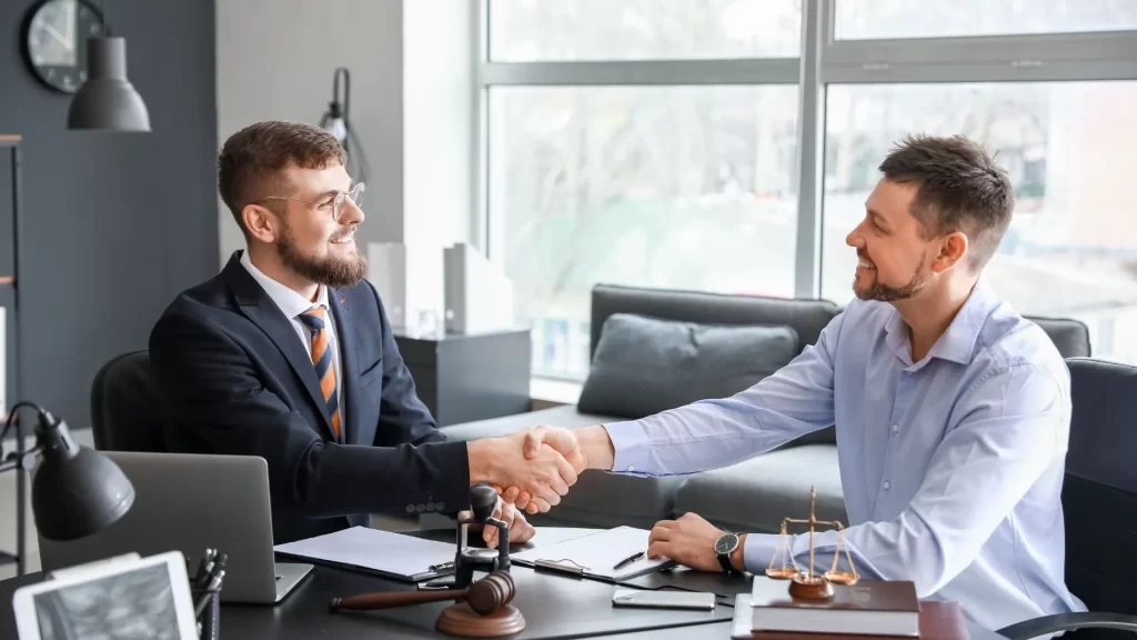 Two men are shaking hands over what seems to be a signed deal. They are both seated in a law office, with one of them looking like a lawyer, and the other another professional who offered their services. 