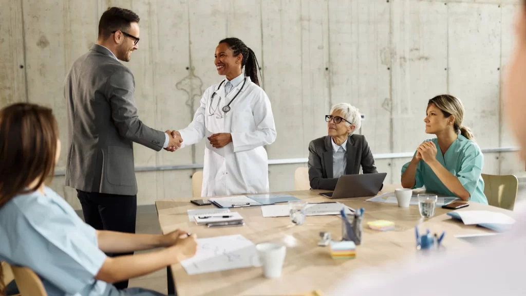 A female doctor is shaking hands with a male business professional as a sign of a closed business deal. The other doctors and managers seem to approve of the closed deal. 