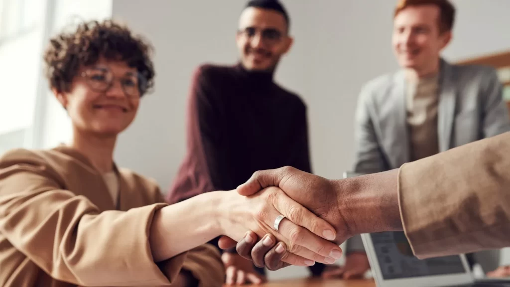 A business hand shake as a sign of a closed deal is in the foreground. In the background are three individuals smiling as the deal is closed. 