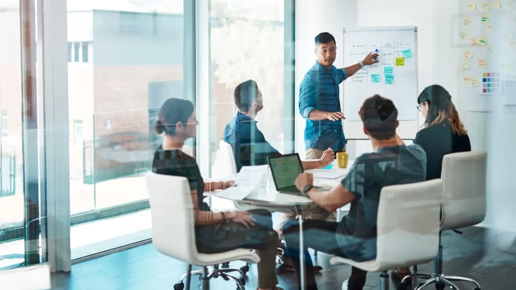 An Asian team leader is discussing something on the office white board to his diverse team. Each team member is listening. 