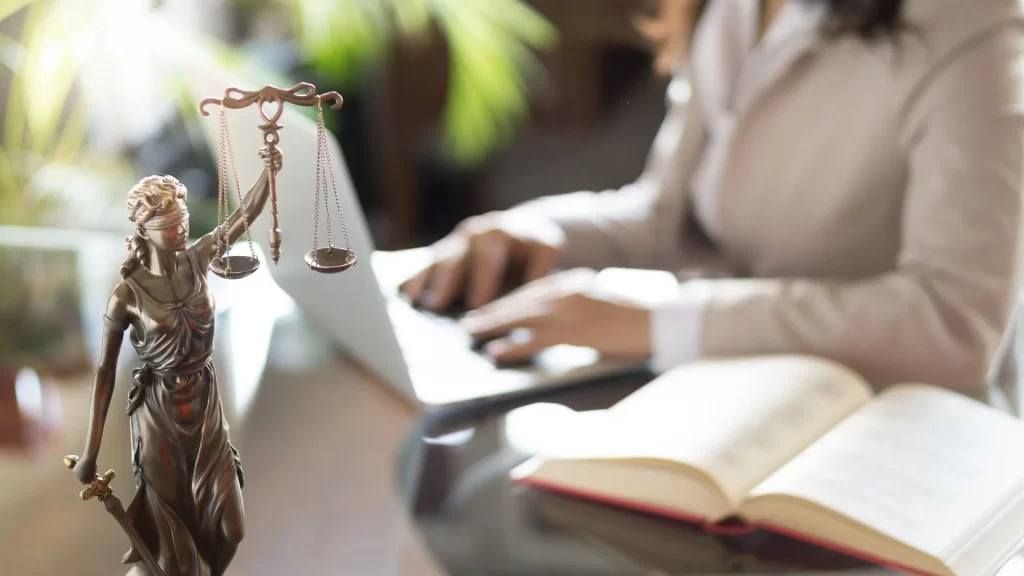 A lady justice figurine is in focus in the photo while a laptop, a typing lawyer, an open law book is in the blurred background 