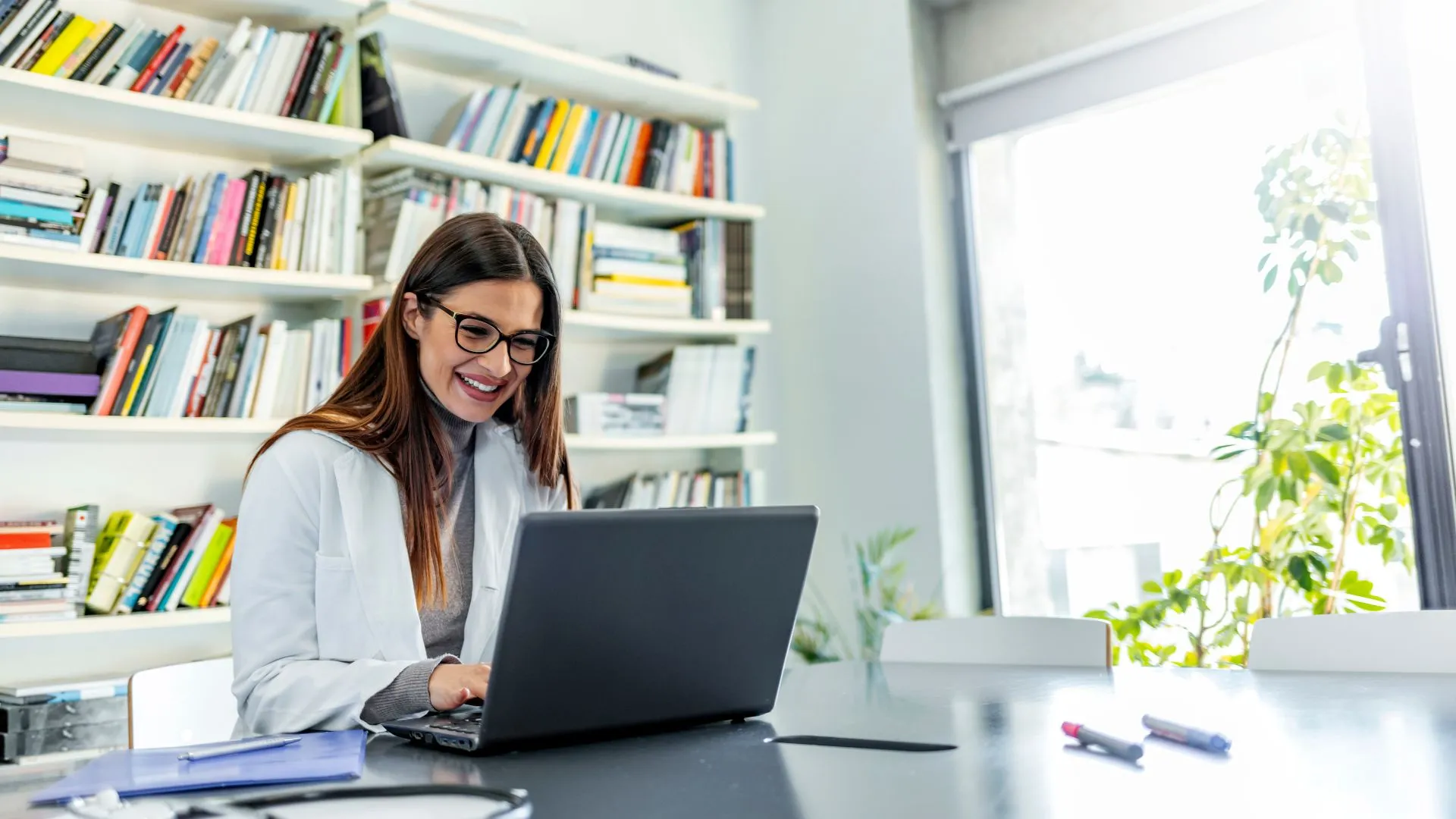 A female doctor is smiling in her office while using her laptop.