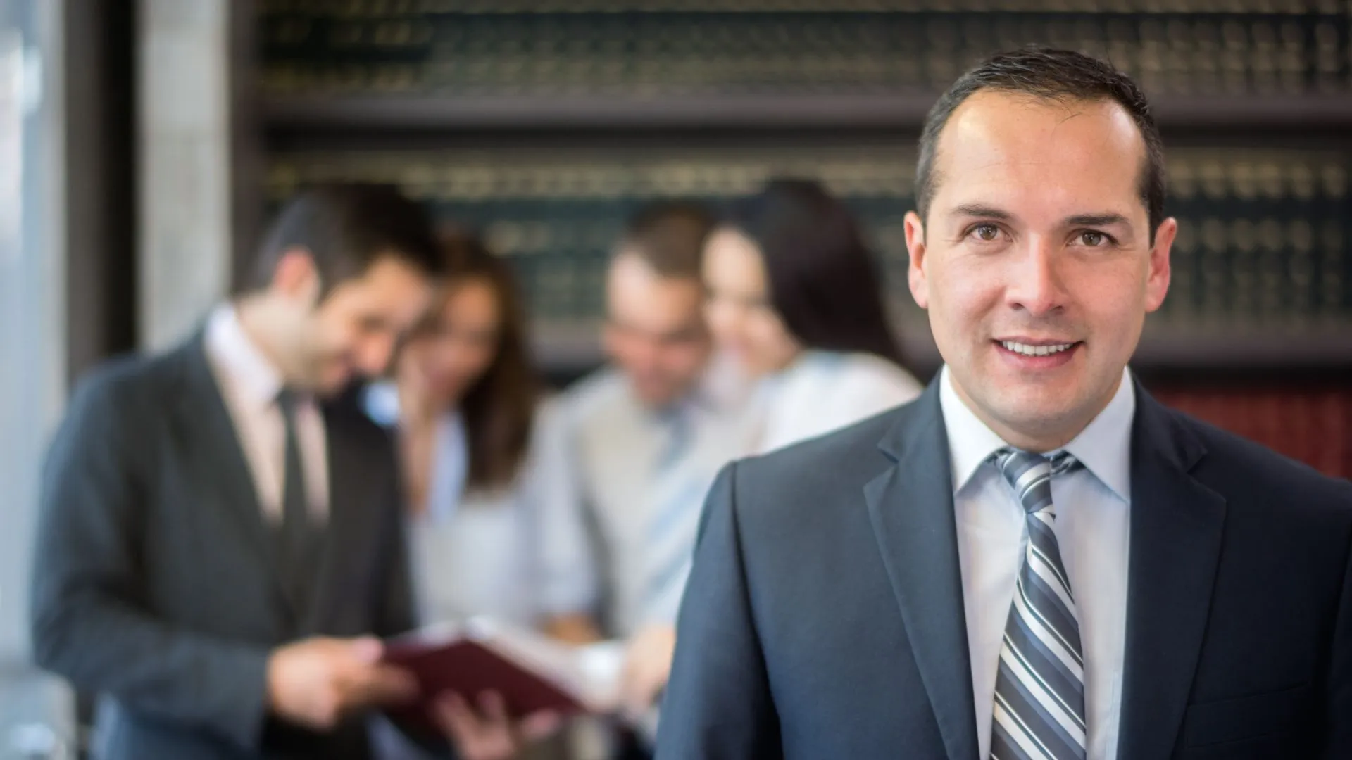 A male lawyer smiling at the camera while his fellow lawyers are discussing something in the background