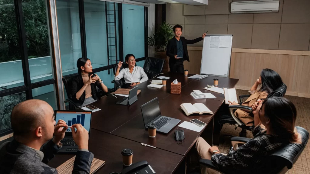 An Asian man is happily presenting something in a small office conference room. His colleagues are listening intently while their laptops and work essentials are all on the table.