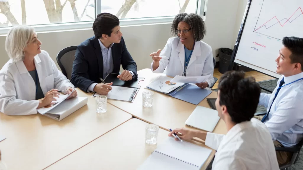 A group of five people comprised of doctors and management professionals seem to be in deep discussion. There is a graph talking about “Expectation” and “sales” on the background of the photo.