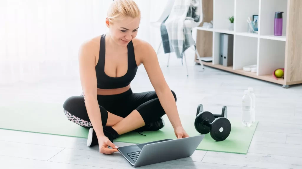 A gym owner in workout clothes is checking her laptop while sitting on a yoga mat