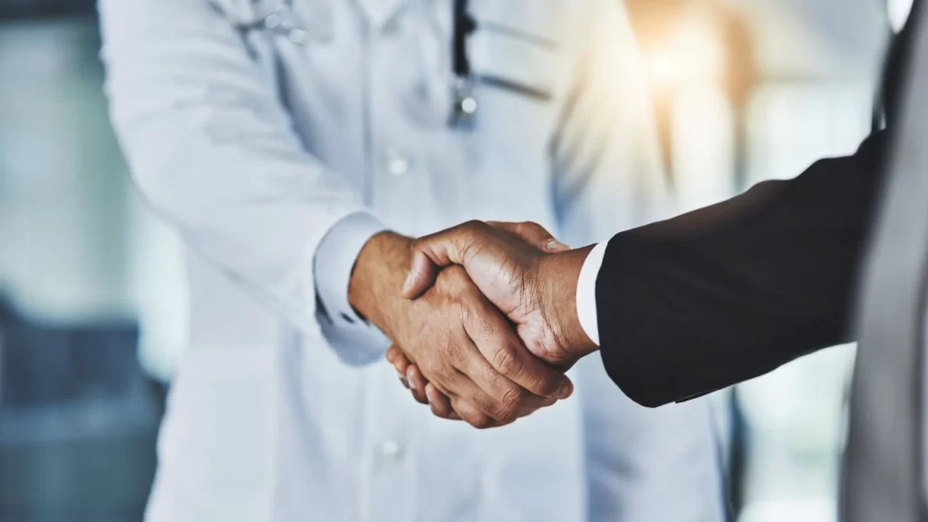 The arm of a doctor is seen to be shaking hands with the arm of an individual in business attire.