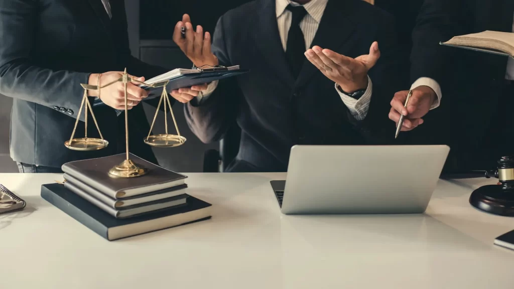 A group of lawyers seem to be in an intent discussion. One hand is seen pointing to a laptop screen.