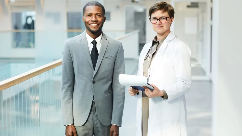 A female doctor is smiling while holding a chart. She is beside a male colleague dressed in business attire who is also smiling.